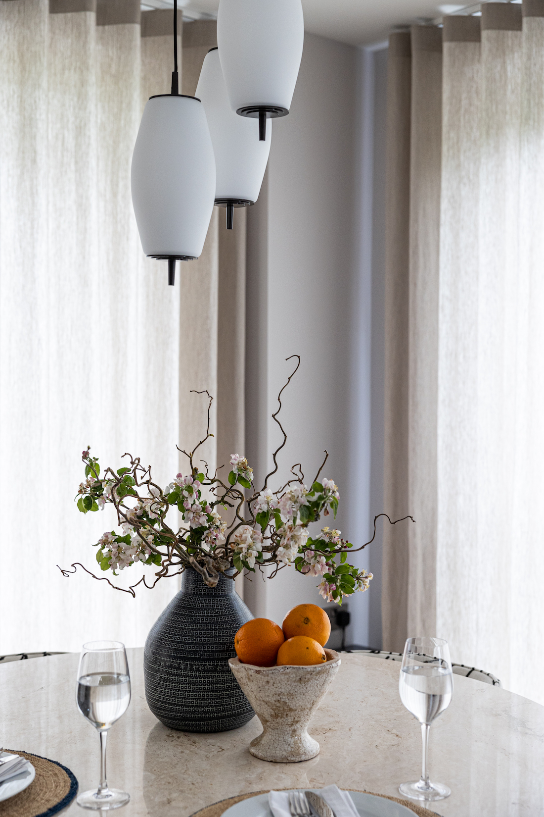 urban contemporary three glad drop pendant lights over a dining table with a bunch of cherry blossoms in a vase, a table setting for two and a bowl of oranges
