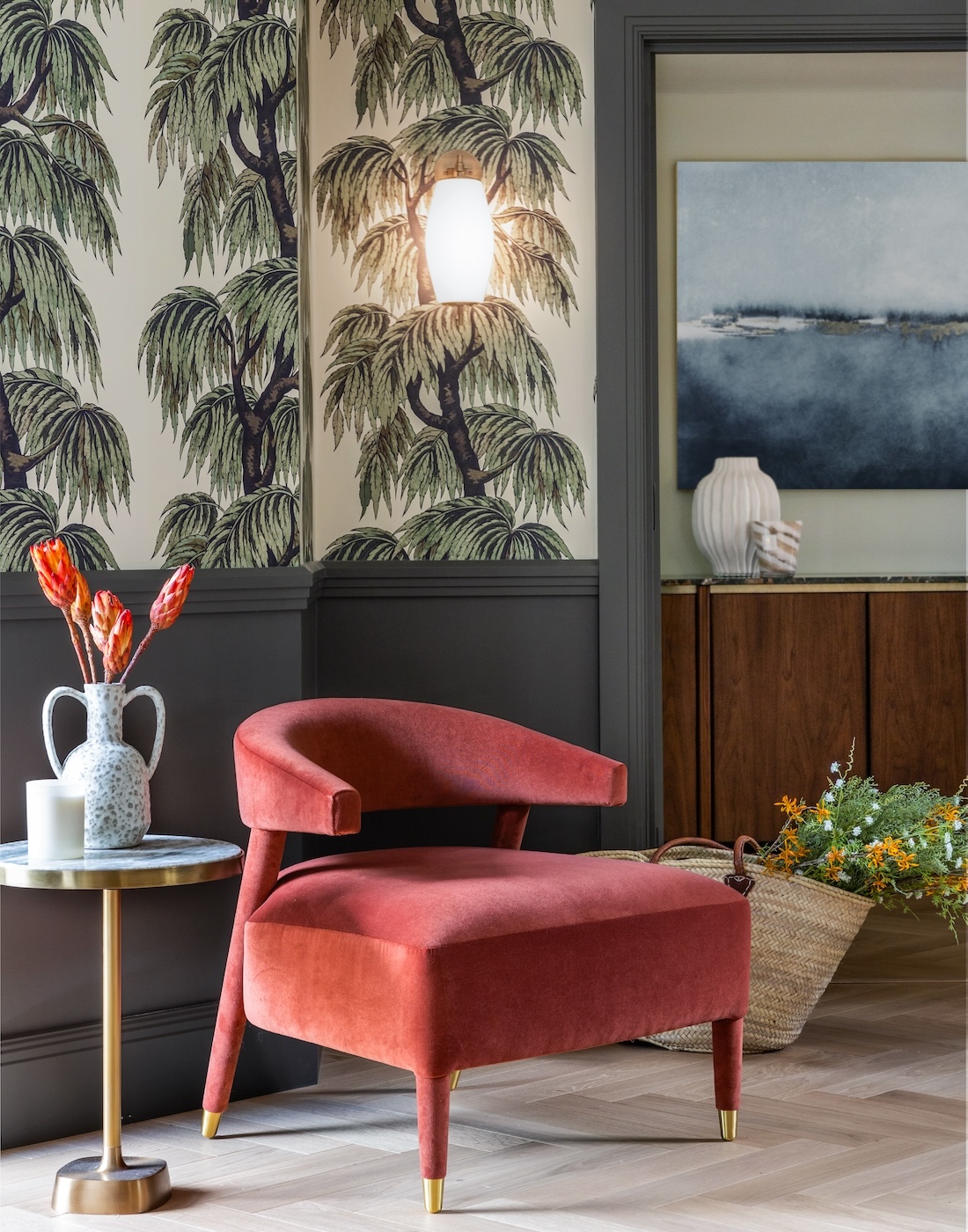 Art Deco Inspired Entrance Hall designed by Pfeiffer Design. Art deco inspired wallpaper, half height panelling and glazed French double doors in deep green. Bronze obscured glass wall lights. A soft red occasion chair with side table with dried flowers in a double handled vase.