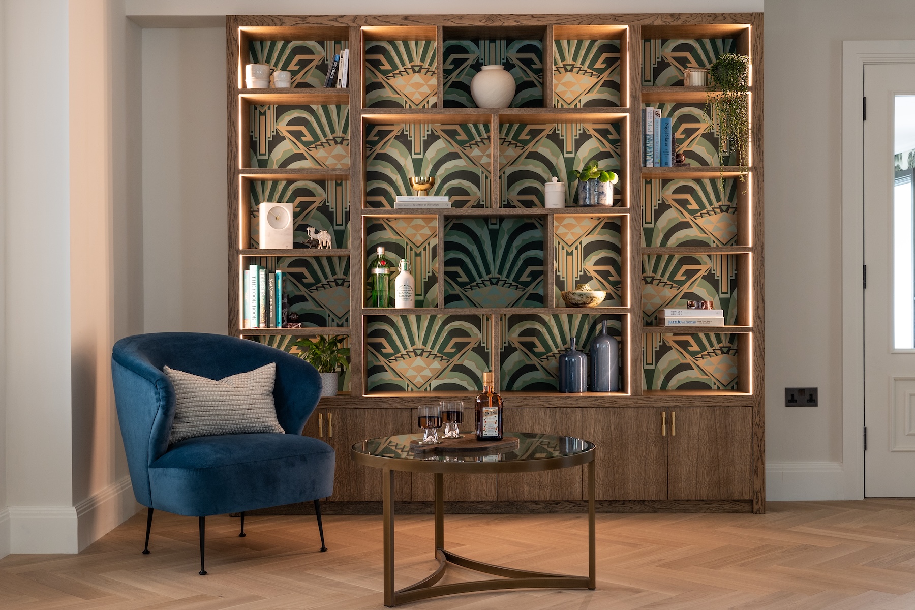 Art Deco Inspired Bespoke Bar and Library made by our bespoke joiners, designed by Pfeiffer Design. Art deco wallpapered insert within light timber open cabinetry, hints of gold through the backing and handles, and LED lighting for a warm but subtle glow