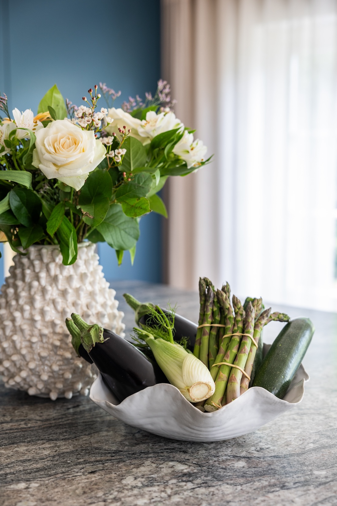 Art Deco Inspired Bespoke Kitchen made by our bespoke joiners, designed by Pfeiffer Design. Bespoke Joinery Kitchen with stone top island. Bunch of flowers in a spiky white ceramic vase and a selection of vegetables in a wavy ceramic white bowl